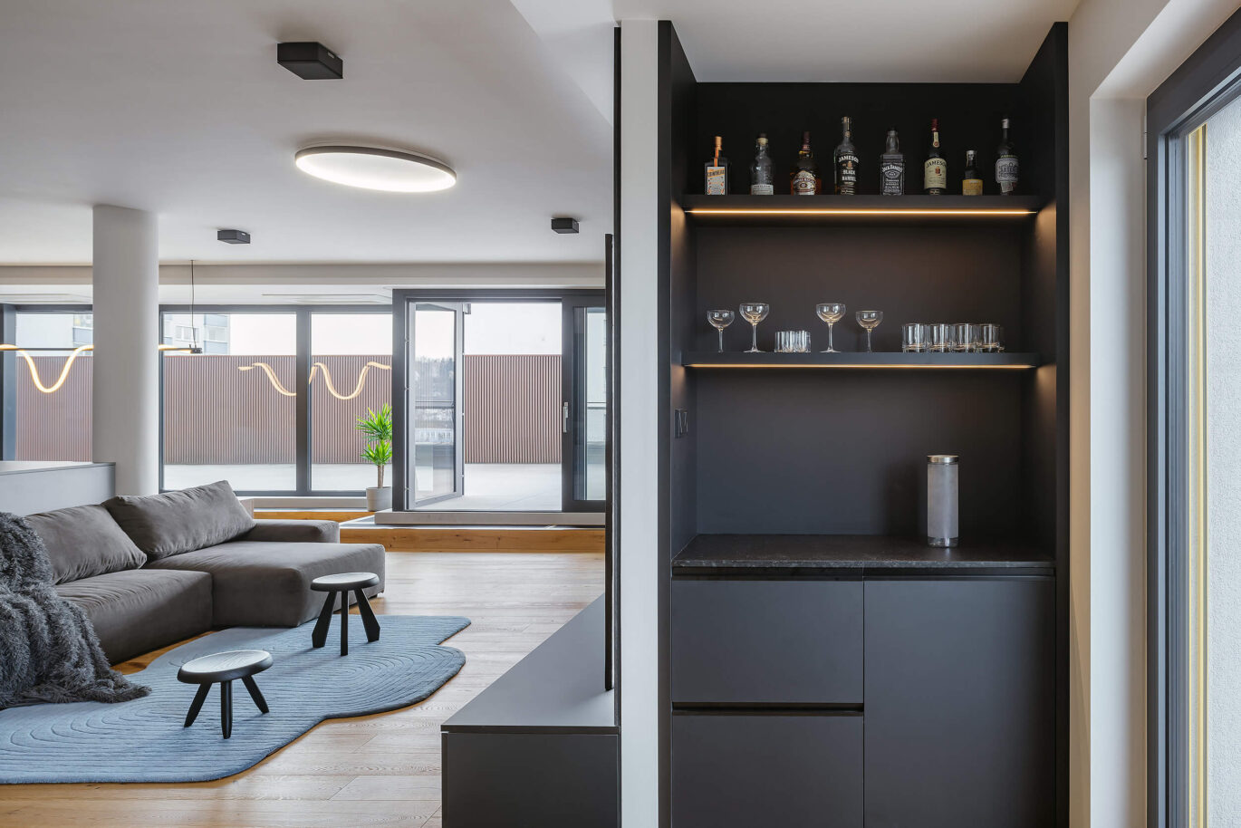 Propojený prostor kuchyně a obývacího pokoje s minimalistickým designem, dřevěnou podlahou, šedými a černými akcenty a integrovaným osvětlením.