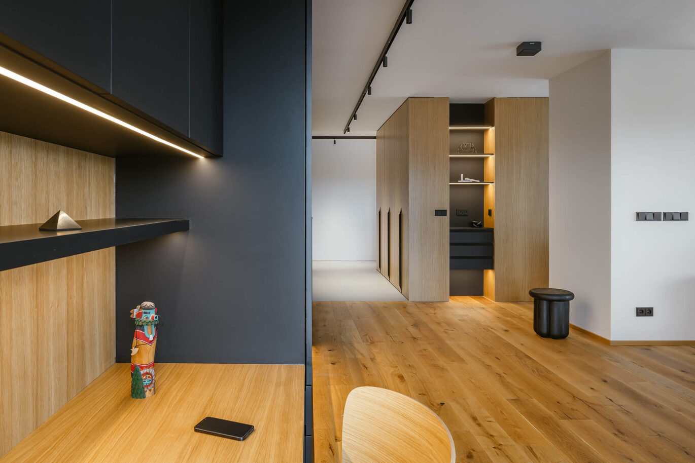 Propojený prostor kuchyně a obývacího pokoje s minimalistickým designem, dřevěnou podlahou, šedými a černými akcenty a integrovaným osvětlením.